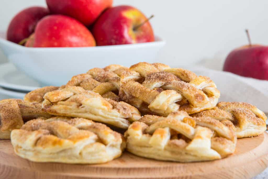 Homemade apple pie cookies with lattice pastry tops and cinnamon sugar, stacked on a wooden board with a bowl of red apples behind them.