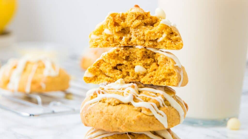 A stack of four soft yellow banana pudding cookies drizzled with white chocolate, the top two broken open to show a crumbly center, with a tall glass of milk and more cookies on a cooling rack behind them.