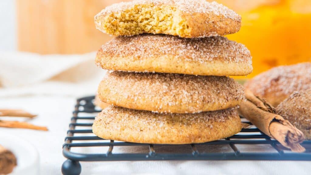 A stack of four homemade pumpkin snickerdoodle cookies coated in cinnamon sugar on a black wire cooling rack, the top one with a bite taken out, with cinnamon sticks beside them.