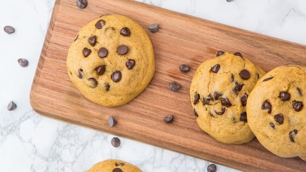 Pumpkin chocolate chip cookies lined up on a wooden board with chocolate chips scattered around them.