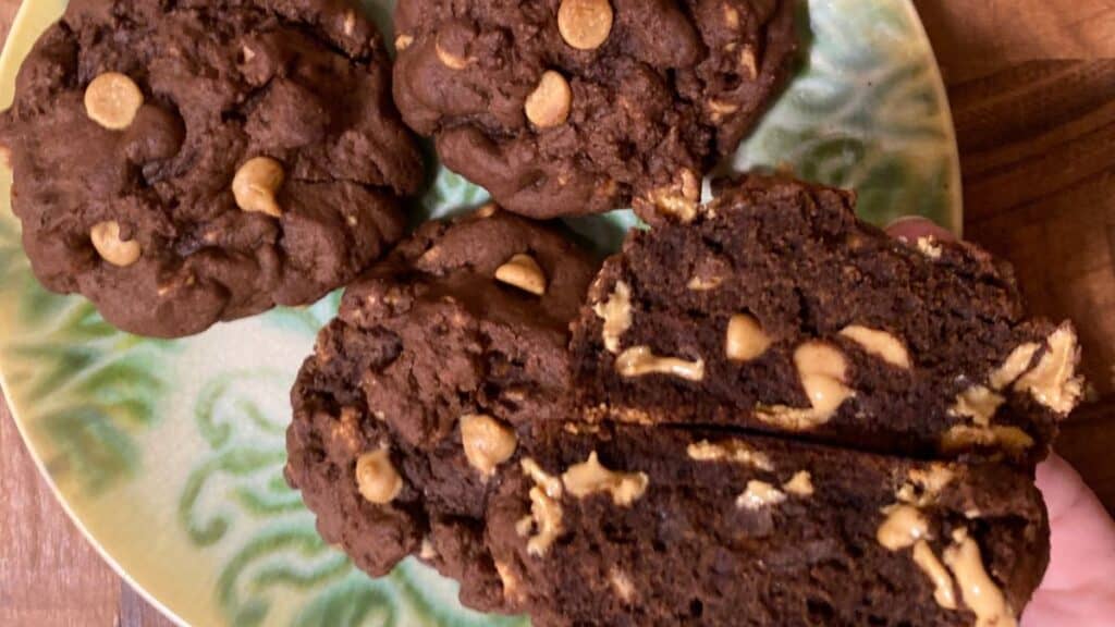 Four homemade dark chocolate peanut butter chip cookies on a green patterned plate, one broken open to show the melted peanut butter chips inside.