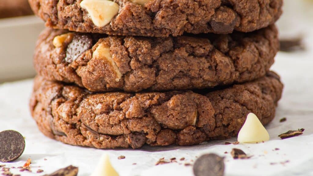 A stack of three homemade fudgy triple chocolate chip cookies studded with white and dark chocolate chips on parchment paper, with chocolate chips scattered around them.