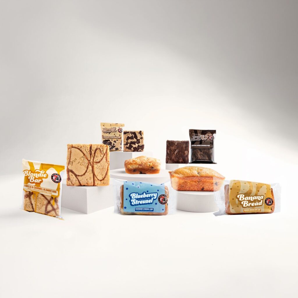 7 Brew's five new bakery items on white pedestals: a Blondie Bar, a Brunette Mocha Brownie Bar, a Chocolate Chunk Cookie Bar, a Banana Bread loaf and a Blueberry Streusel loaf, each shown wrapped and unwrapped.