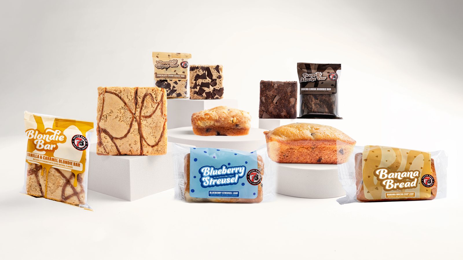 7 Brew's five new bakery items on white pedestals: a Blondie Bar, a Brunette Mocha Brownie Bar, a Chocolate Chunk Cookie Bar, a Banana Bread loaf and a Blueberry Streusel loaf, each shown wrapped and unwrapped.