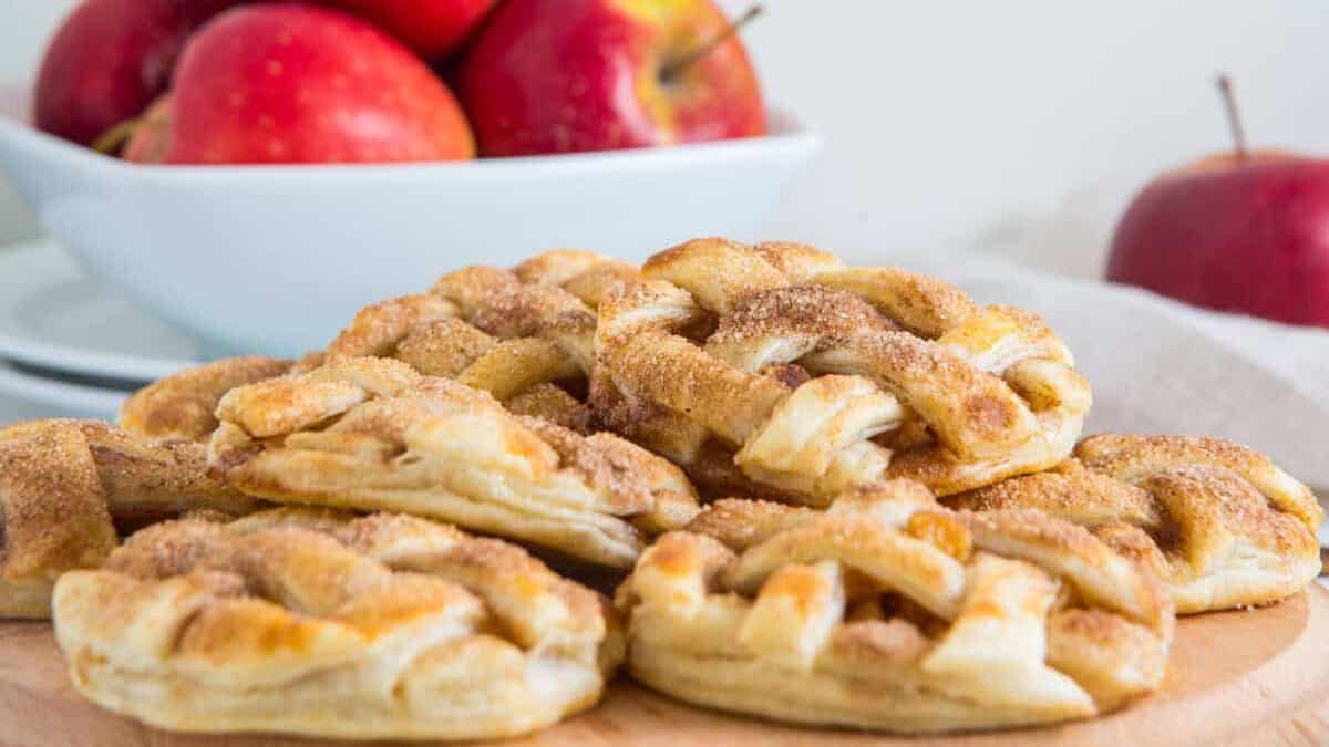 Lattice-topped apple pie cookies made with flaky pie crust and dusted with cinnamon sugar, stacked on a wooden board in front of a bowl of red apples.