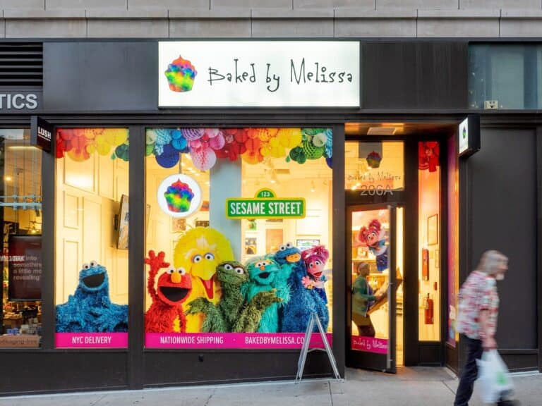 The storefront of a Baked by Melissa cupcake shop in Manhattan, with a colorful window display and a person walking past.