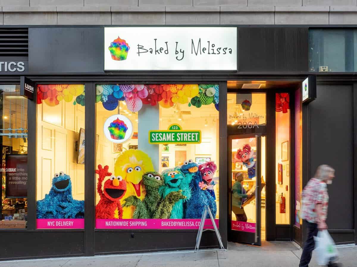 The storefront of a Baked by Melissa cupcake shop in Manhattan, with a colorful window display and a person walking past.