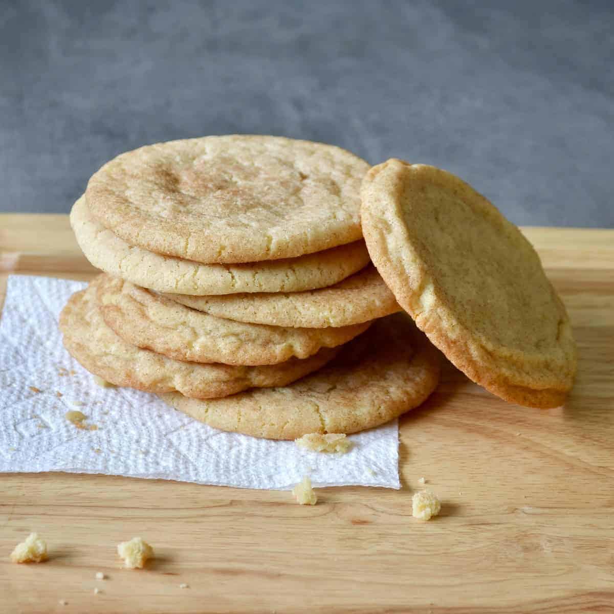 A stack of pale, flat snickerdoodle cookies dusted with cinnamon sugar on a paper towel and wooden board, with one cookie leaning against the stack.