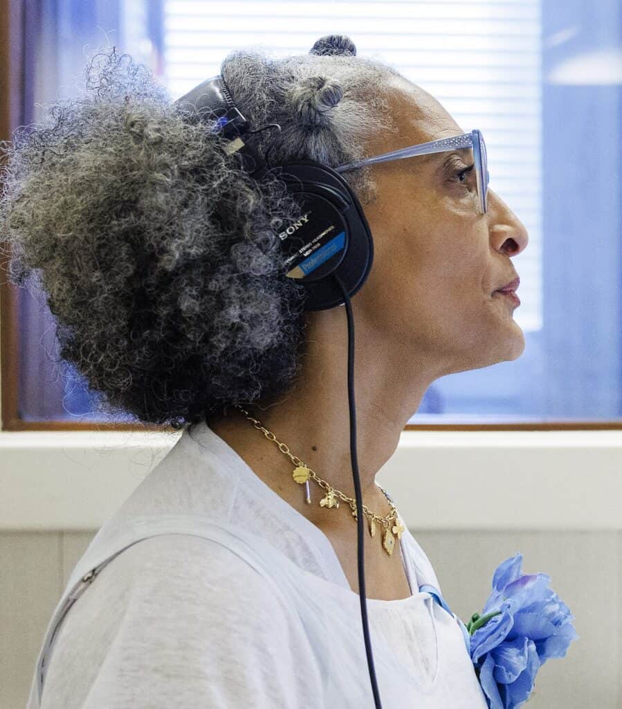 Carla Hall, in profile with silver curls, light blue glasses and studio headphones, during a recording session.