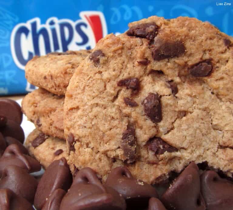 Close-up of Chips Ahoy! chocolate chip cookies leaning in a stack, surrounded by loose chocolate chips, with a blue Chips Ahoy! package behind them.