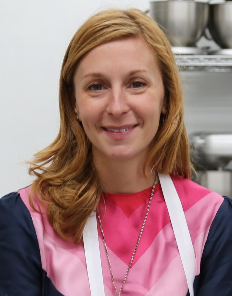 Christina Tosi, smiling, with shoulder-length strawberry-blond hair, wearing a pink chevron top and a white apron in a kitchen with steel mixing bowls on a shelf behind her.