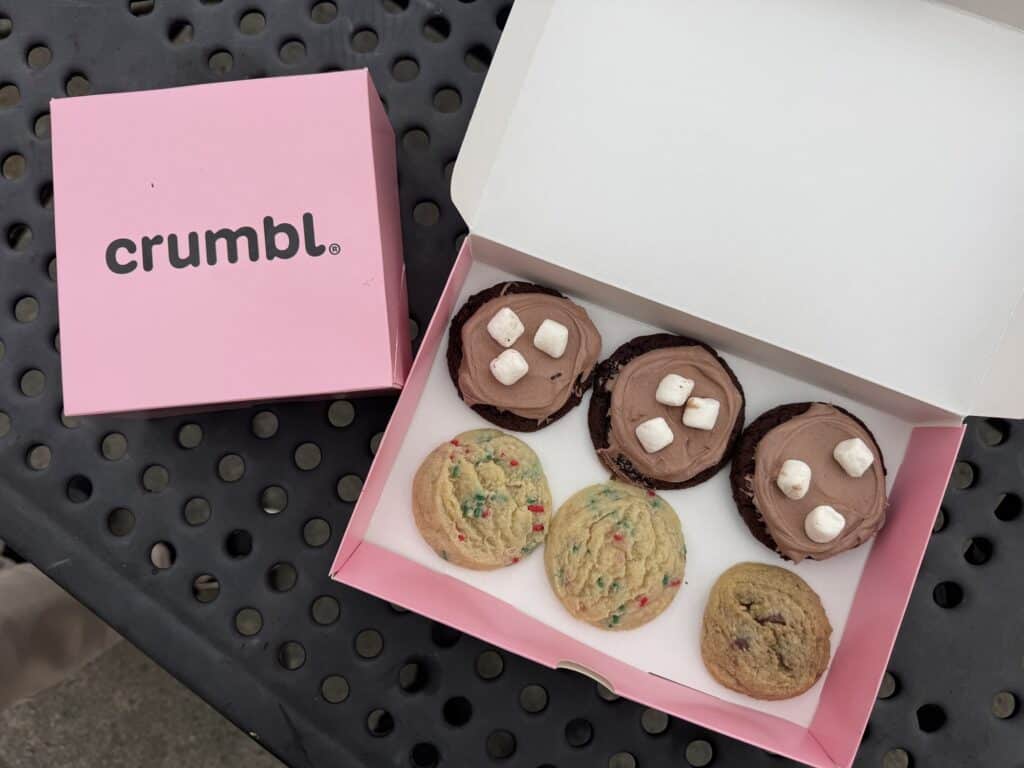An open pink Crumbl box holding six cookies, three chocolate with marshmallows and three with sprinkles, beside a closed pink Crumbl box.