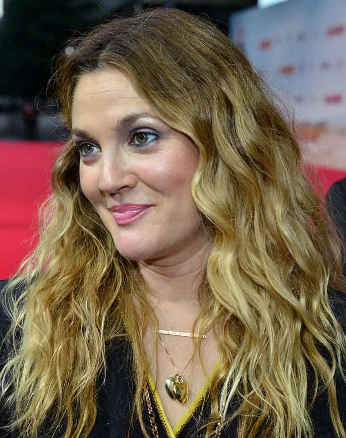 Drew Barrymore, with long wavy blonde hair and a gold heart necklace, smiles on a red carpet.
