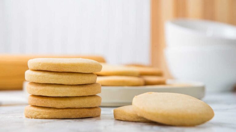 A stack of five round golden shortbread cookies beside a plate of more shortbread on a marble counter.