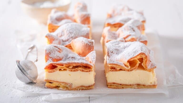 Squares of karpatka, a Polish cake of puffed choux pastry layered with thick vanilla cream, dusted with powdered sugar beside a small sieve.