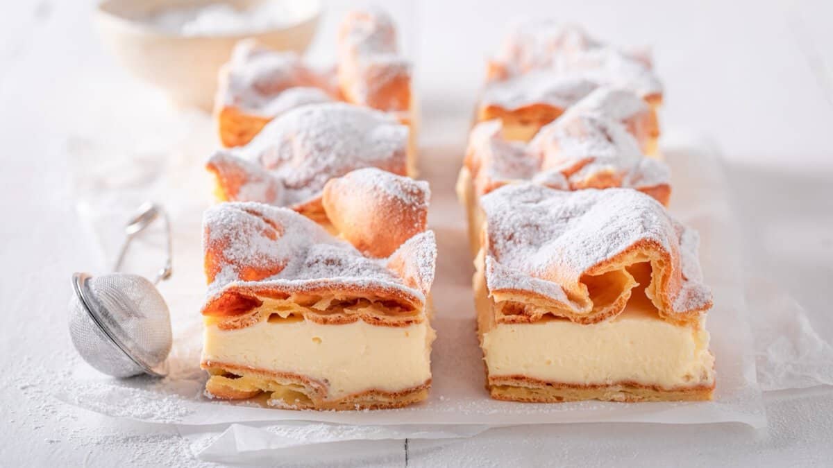 Squares of karpatka, a Polish cake of puffed choux pastry layered with thick vanilla cream, dusted with powdered sugar beside a small sieve.