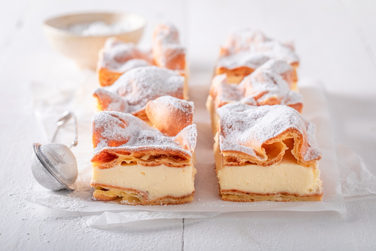 Squares of karpatka, a Polish cake of puffed choux pastry layered with thick vanilla cream, dusted with powdered sugar beside a small sieve.