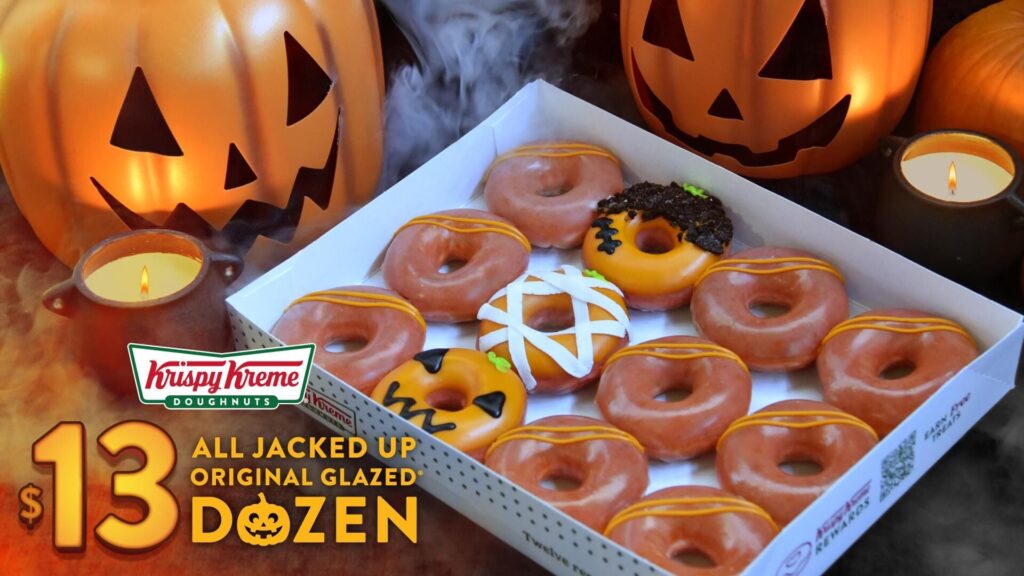 Krispy Kreme's promotional image for its $13 All Jacked Up Dozen: an open box of orange-glazed doughnuts, including a mummy, a jack-o'-lantern and a monster doughnut, beside lit jack-o'-lanterns and a candle.