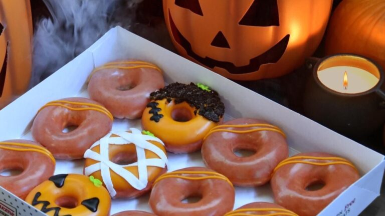 An open box of Krispy Kreme's orange-glazed All Jacked Up Dozen doughnuts, with a mummy doughnut, a jack-o'-lantern doughnut and a monster doughnut topped with chocolate cookie pieces, beside a lit jack-o'-lantern.