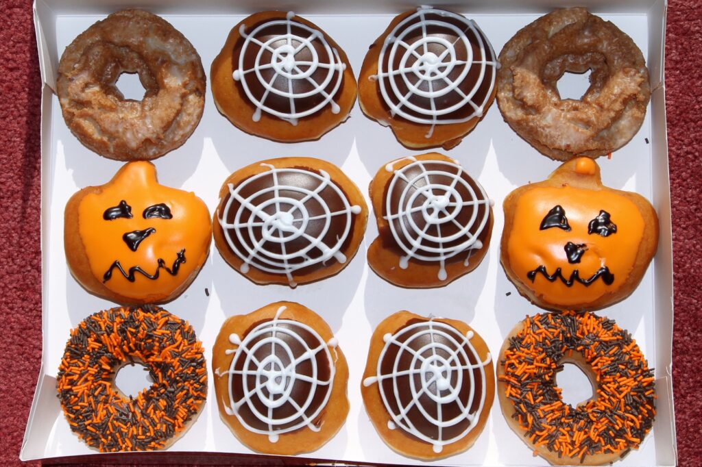 An open box of twelve Krispy Kreme Halloween doughnuts from 2012: two glazed cake doughnuts, six chocolate-iced doughnuts piped with white spiderwebs, two orange jack-o'-lantern doughnuts with black icing faces, and two with orange and brown sprinkles.