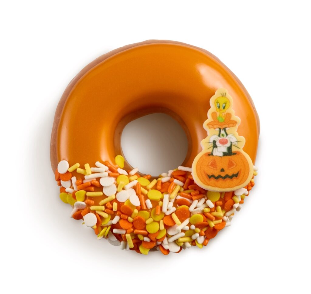 A ring doughnut dipped in orange candy corn flavored icing, with yellow, orange and white confetti sprinkles along the bottom edge and a sugar piece of Tweety and Sylvester sitting on a jack-o'-lantern.
