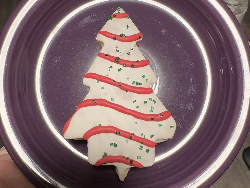 A Little Debbie Christmas Tree Cake with white icing, red icing stripes and green sugar sprinkles, on a purple plate.