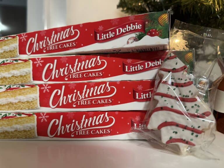 Four stacked boxes of Little Debbie Christmas Tree Cakes with one wrapped tree-shaped cake, white icing with red stripes and green sugar, leaning against them.