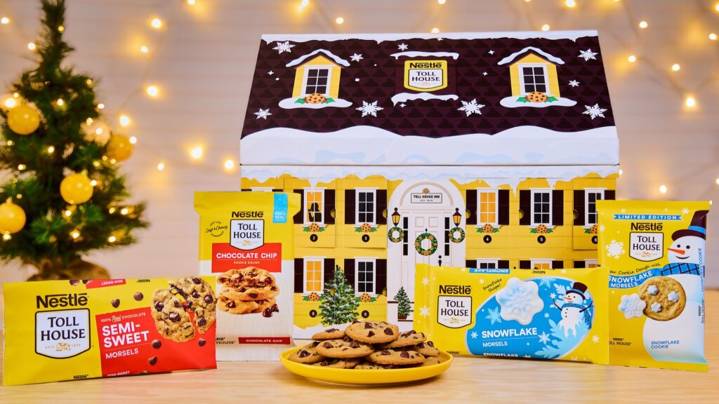 A yellow house-shaped Nestlé Toll House Advent calendar box with a snowy brown roof and numbered doors, surrounded by bags of Semi-Sweet Morsels and Snowflake Morsels, packages of Chocolate Chip and Snowflake Cookie Dough, and a plate of chocolate chip cookies, with a lit Christmas tree behind.