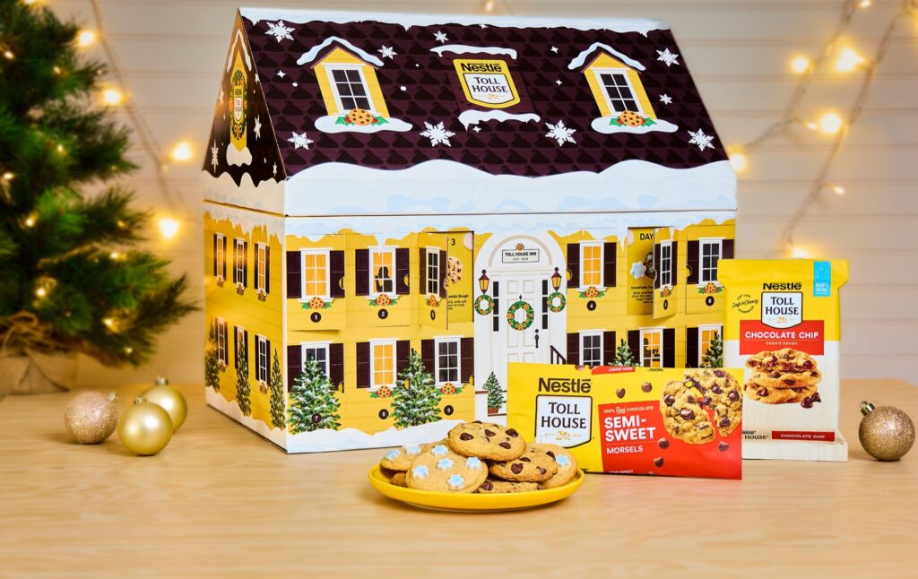 A side view of the house-shaped Nestlé Toll House Advent calendar showing its numbered doors, beside a bag of Semi-Sweet Morsels, a package of Chocolate Chip Cookie Dough, a plate of cookies and gold ornaments.