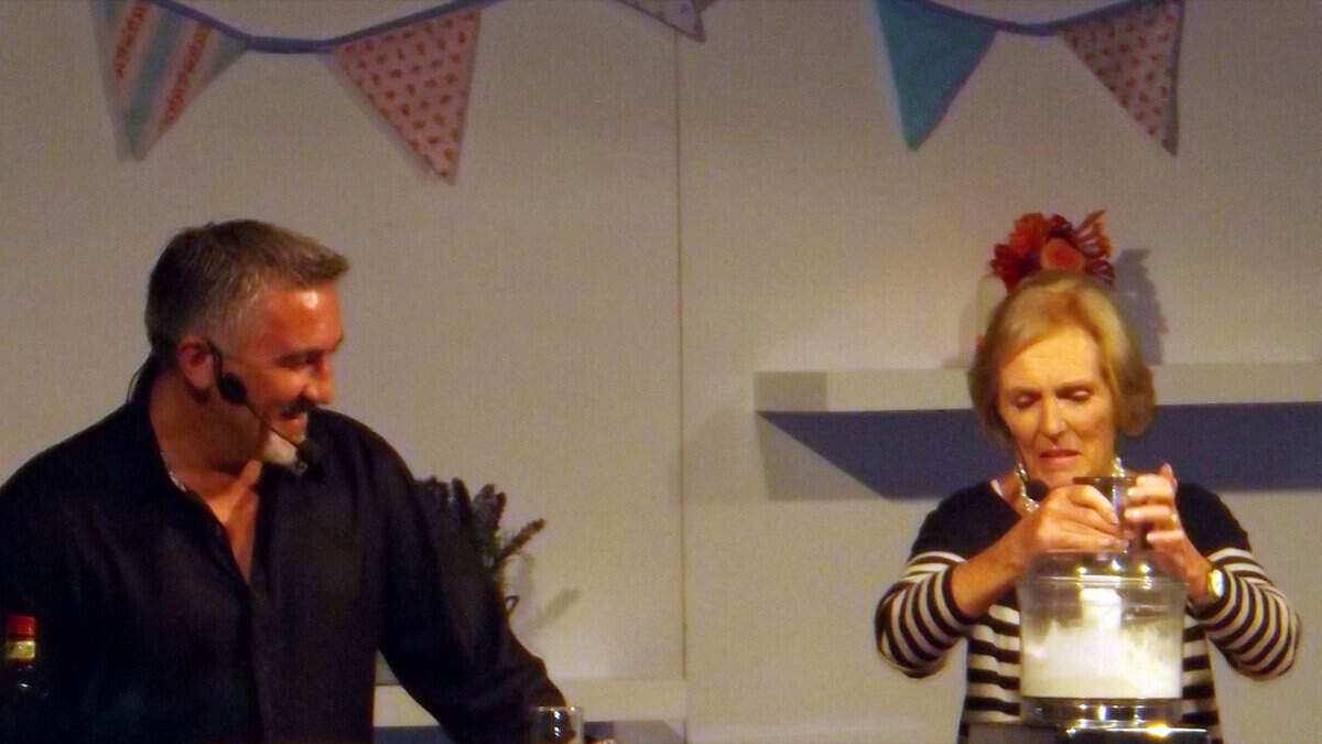 Paul Hollywood, in a black shirt and headset microphone, smiles across a stage at Mary Berry as she works a food processor under colorful bunting.