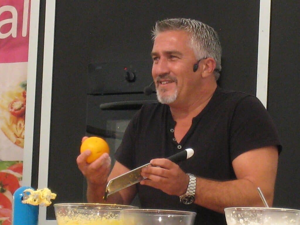 Paul Hollywood, smiling in a black shirt with a headset microphone, holds an orange and a zester over mixing bowls during a cooking demonstration.