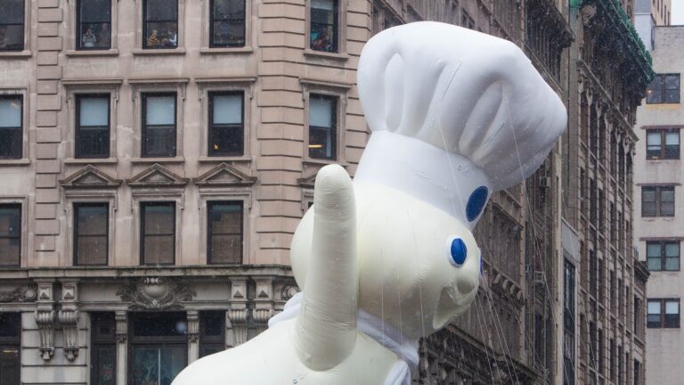 The giant Pillsbury Doughboy balloon in his white chef's hat floats between New York buildings during the 2014 Macy's Thanksgiving Day Parade, one hand raised in a wave.