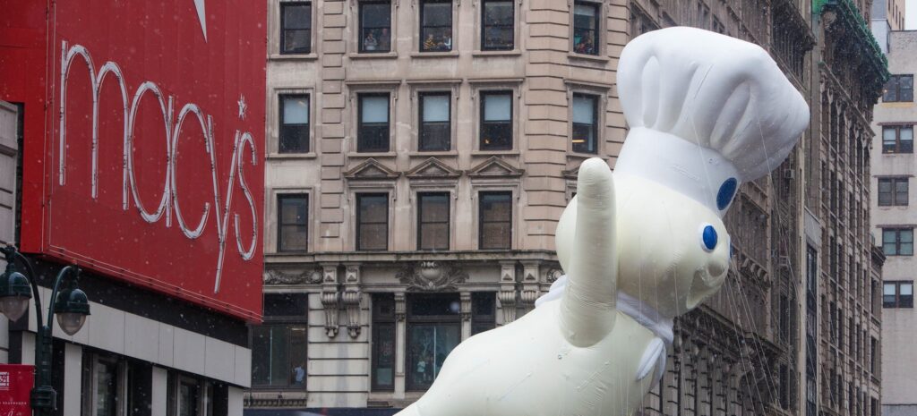 The giant Pillsbury Doughboy balloon, in his white chef's hat and neckerchief, waves as he floats past the red Macy's sign on a snowy New York street during the 2014 Macy's Thanksgiving Day Parade.