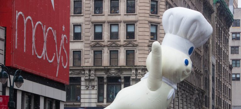 The giant Pillsbury Doughboy balloon, in his white chef's hat and neckerchief, waves as he floats past the red Macy's sign on a snowy New York street during the 2014 Macy's Thanksgiving Day Parade.