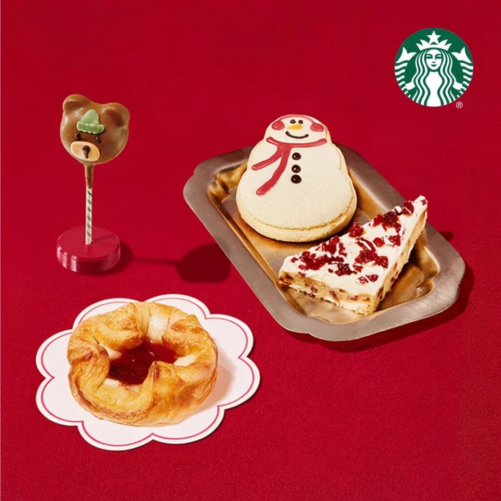 Starbucks' Snowman Cookie, a slice of Cranberry Bliss Bar and a Sugar Plum Cheese Danish on a red background, with the Bearista Cake Pop standing beside them.