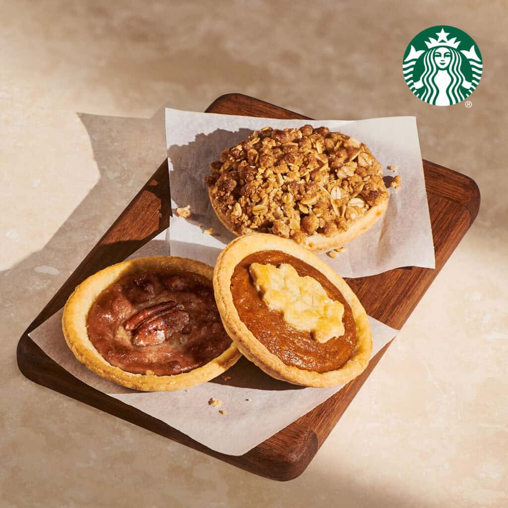 Three Starbucks Mini Pies, apple crumble, pecan and pumpkin, on parchment paper on a wooden board.