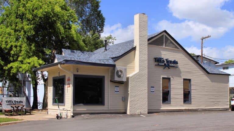 The Tiff's Treats store in central Austin, Texas, a cream-colored converted 1940s bungalow with a brick chimney, a blue Tiff's Treats cookie delivery sign on its gable, a large front window and a picnic table under a tree, photographed in July 2022.