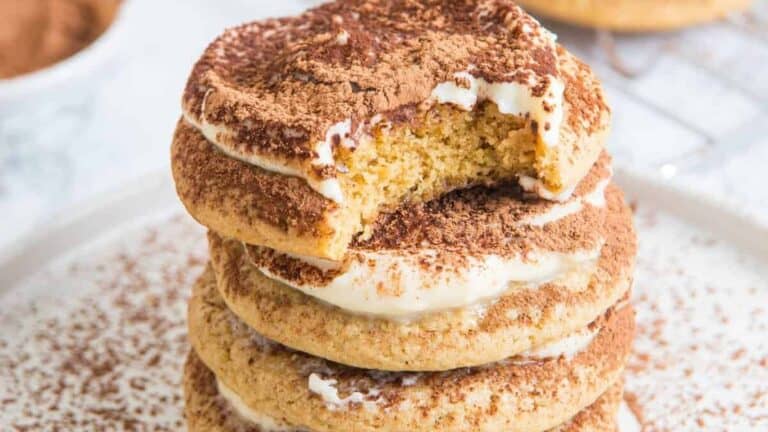 A stack of tiramisu cookies dusted with cocoa, with mascarpone frosting between the layers and a bite taken from the top cookie.