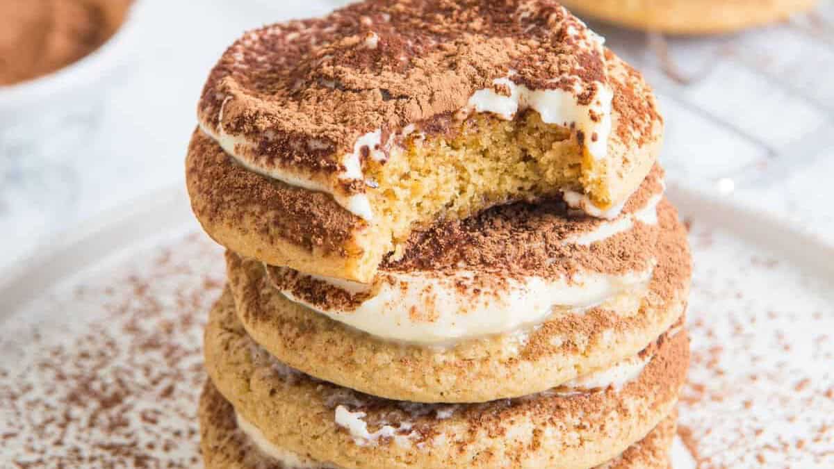 A stack of tiramisu cookies dusted with cocoa, with mascarpone frosting between the layers and a bite taken from the top cookie.