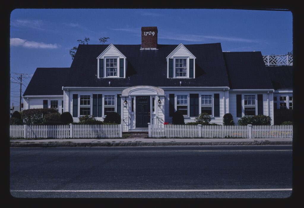 A white Cape-style house with black shutters, two dormer windows, a brick chimney and a white picket fence, seen from across a two-lane road.