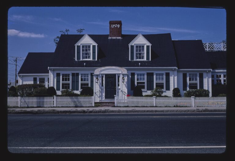 A white Cape-style house with black shutters, two dormer windows, a brick chimney and a white picket fence, seen from across a two-lane road.
